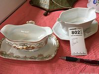 Two gravy boats photographed together on a surface with a pen for scale, showing one white six-sided boat with detached underplate and one floral painted Limoges boat with attached underplate.