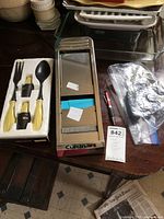 Photo showing the Cuisinart harp slicer next to molded white foam holding yellow-handled OXO kitchen tools including a peeler, placing fork, and a small knife, on a dark wooden table along with stacked white ceramic serving dishes.