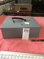 Gray insulated lock box measuring 12 x 10 x 4.5 inches with a black handle and key on a pink surface next to a keyboard and pen.