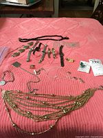 Top view of six wristwatches with various straps, many metal, alongside multiple necklaces, bracelets, and earrings laid out on a pink textured cloth.