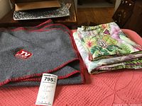 Gray fleece lap throw with red stitching and TV Guide logo beside folded leaf motif bedding sheets and pillowcases