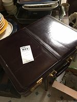 Dark brown leather briefcase closed with visible metal combination locks and a buyer sticker on the front.