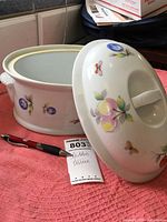 Oval ceramic tureen with lid, showing fruit motif decoration and side handles, placed on pink towel with pen and label beside.