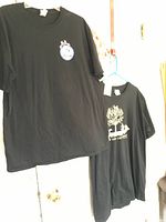Photo showing two black T-shirts hanging on hangers: one with a crab logo (Jimmys Famous Seafood) on the upper left chest, the other with a Dog is Good graphic featuring a tree and two dogs.