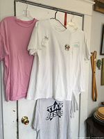 Four XL size T-shirts hanging on hangers against a white door. Pink plain tee, white tee with Maryland crab and Buddy by the Sea design, white tee with teal text design, and gray Team Jesus tee partially folded.