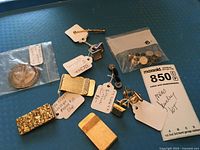 Photo showing various men's jewelry items on blue background, including money clips, cuff links, tie tacs, a souvenir coin in a plastic pouch, and price tags.