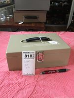 Top and front view of beige metal lock box with black handle and silver latch, placed on a pink textured cloth with a pen for scale.