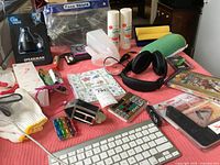 Overview of miscellaneous household and office items on a red table cover including gloves, face shield, markers, headphones, calculator, pepper spray, disposable gloves, and paper products.