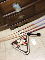 Photo showing four pool cues, a brush, a triangle rack, and a set of pool balls including cue ball and numbered balls with solids and stripes.