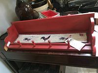 Photo showing the entire red coat rack lying flat among other items. Distressed red paint, four hooks, long wooden piece with slightly raised edges and rooster motif in the center panel.