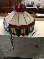 Stained glass shade with red, white and green squares and rectangles arranged in a pattern. The top has a smaller flared section with red glass. The shade is placed on a white surface with a chain hanging down.
