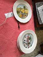 Top view showing the top plate with a colorful, detailed Grandma Moses style rural scene on a white ceramic plate with scalloped edges. Plates are stacked with similar designs.