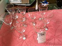 Ten clear small wine glasses by Lenox with gold rims arranged on a pink textured surface, all upright and visible from the top and side.
