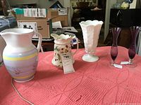 Lot shown on pink textured cloth including two purple bud vases, white milk glass grape vase, and two ceramic pitchers with strawberry and grape motifs.