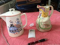 Two pottery pitchers on a pink textured cloth surface, one lidded Canonsburg Pottery pitcher with girl motifs and one Little Bo Peep shaped pitcher.