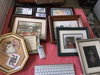 Photo showing multiple framed artworks laid out on a bed or surface, with notable pieces including a watercolor landscape and an octagonal framed decorative art with an angel figure and playing card.