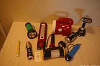 Image of 10 assorted flashlights, a red multi-function radio with handle, and a windup device, placed on a beige surface under bright lighting.