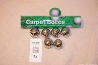 Six metal bocce balls arranged in front of green container tube labeled 'Carpet Bocce' on a beige surface.