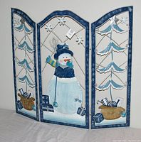 Front view of three-panel Christmas fireplace screen showing central snowman and decorated side panels with Christmas trees and gift baskets.