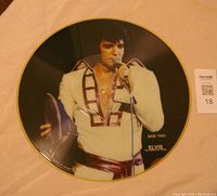 Front side of the vinyl LP picture disc showing Elvis Presley in a white jumpsuit performing with microphone and tambourine.