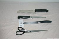 Four Henkel kitchen cutting tools displayed on light fabric background, including three knives and one pair of scissors.