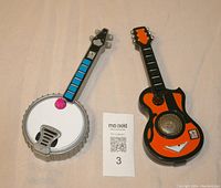 Front view of two micro jammer toys shaped like banjo and guitar, with a small note showing lot number.