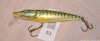 Photo showing entire muskie fishing lure with attached hooks and diving lip, placed on a flat surface with a tag beneath it.