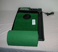 Electric putting green system black base with green felt mat partially rolled, showing hole and ball return area, power cord attached.