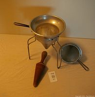 Three items arranged on a table: a metal fruit strainer with integrated fine mesh basket and tripod stand, a second similar metal strainer, and a solid wooden mallet with a wide pressing end and rounded handle.