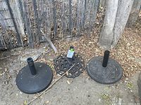 Three black metal umbrella stands showing different base designs and wear, placed outdoors on ground with dry leaves.