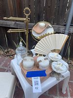 Full view of bathroom vanity items on a small white table outside, showing brass towel holder with round mirror, glass jar, candles with decorative holders, soap dishes, and a folding hand fan.