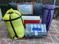 Camping gear items arranged together showing cooler, tent bag, sleeping bag, sleeping pad, and sand mat.
