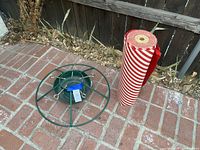 Green metal Christmas tree stand with multiple adjustable eye bolts and central base, shown on brick ground next to a large roll of gift wrap.