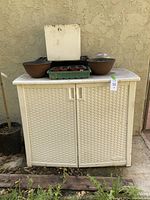 Full view of white plastic Suncast garden storage cabinet with lid closed and assorted planters on top including ceramic bowls, seed starter trays, and a white ceramic planter