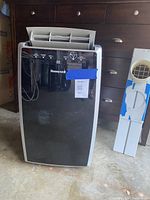 Front view of black and silver Honeywell portable air conditioner with control panel and display visible. Next to it is the partial window attachment kit with round vent.