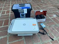 Shows HP DeskJet 2755e printer, black desk chair wheel set in open box, blue and black plastic file box