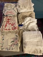 Overall view of folded assorted table linens showing embroidered, lace, and quilted pieces in white, cream, and multicolor designs.