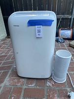 Front view of white Toshiba portable air conditioner with attached exhaust hose next to unit, outdoors on brick surface.