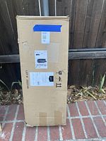 Tall cardboard box with shipping labels on a brick floor, showing original packaging condition and shipment details.