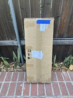 Sealed upright cardboard package containing the garden planter box, standing against a wooden fence outdoors, showing shipping labels and handling symbols.