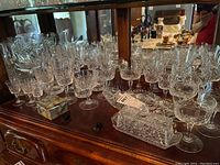 Overall view of glassware collection on a wooden surface in front of mirror, showing multiple cut pressed glasses of varying shapes and sizes, butter dish, and pickle dish with lid.