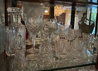 Full view of decanter, wine glasses, bells, and bud vase arranged on a glass cabinet shelf with reflective background.