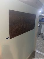 Photo showing the full carved wooden art panel mounted on a wall, rectangular shape, dark wood with visible cultural carvings.