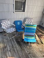 Four outdoor chairs positioned on wooden deck: two aluminum lawn chairs with woven straps, one blue folding Prima Jack chair, one blue-green striped beach chair with wooden armrests.