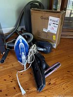 Photo showing a T-Fal iron, Miele Classic C1 vacuum with hose, wand, and floor brush attachment, and a cardboard box for Grelife box fan.