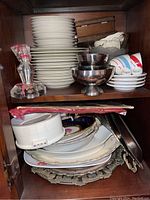 View of stacked Mikasa dinner plates, metal bowls, teacups with saucers, glass bowls, and candle holder arranged on wooden shelving unit.