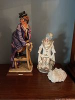 Front view of three items on wooden surface against blue wall: large G. Armani clown figurine, smaller ceramic clown figurine with dogs, and clear crystal rock.