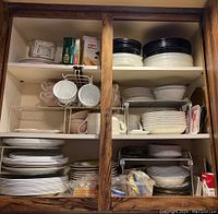Three wooden shelves filled with stacked Corelle dishware including plates, bowls, cups, and mugs in mostly white and black-rimmed designs.