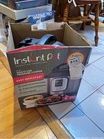 Box of Instant Pot Duo 60 V4 multi-use pressure cooker with graphics and model information visible, placed on floor near a wooden edge.