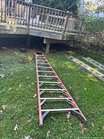 Whole ladder length showing red side rails, metal steps and supports laid flat on grass outdoors